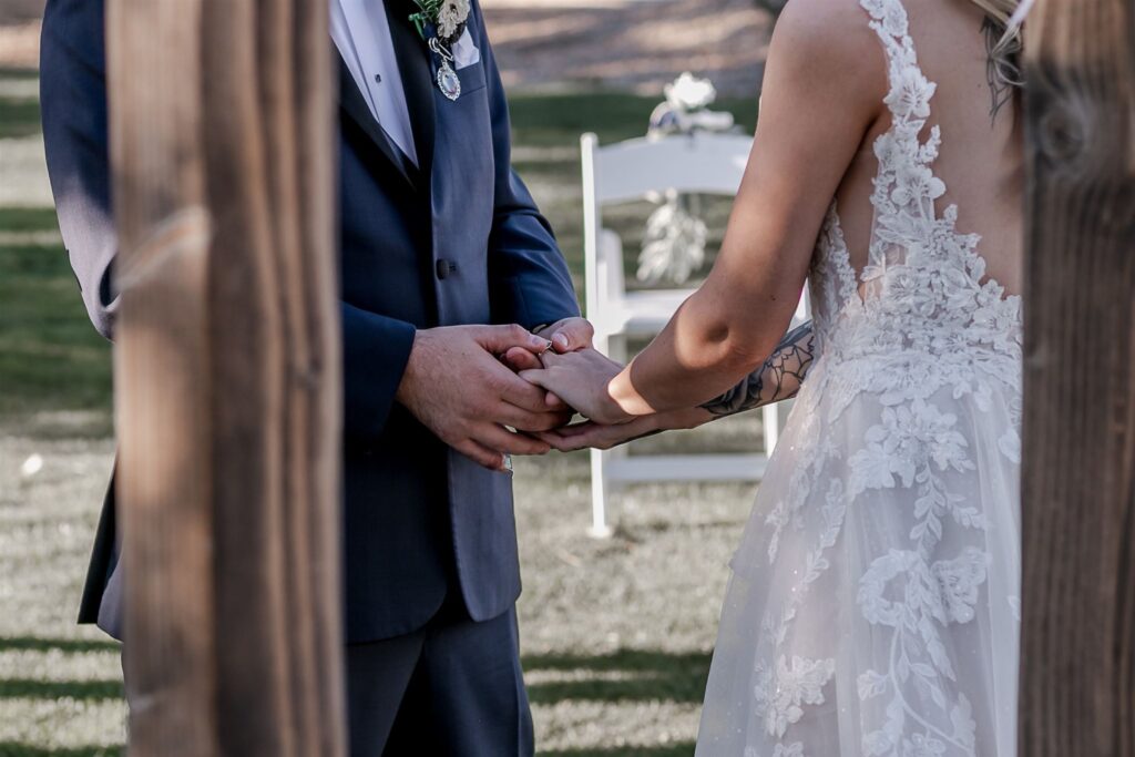 Bride in a white floral lace dress and groom in a dark suit holding hands while placing a ring on her finger.