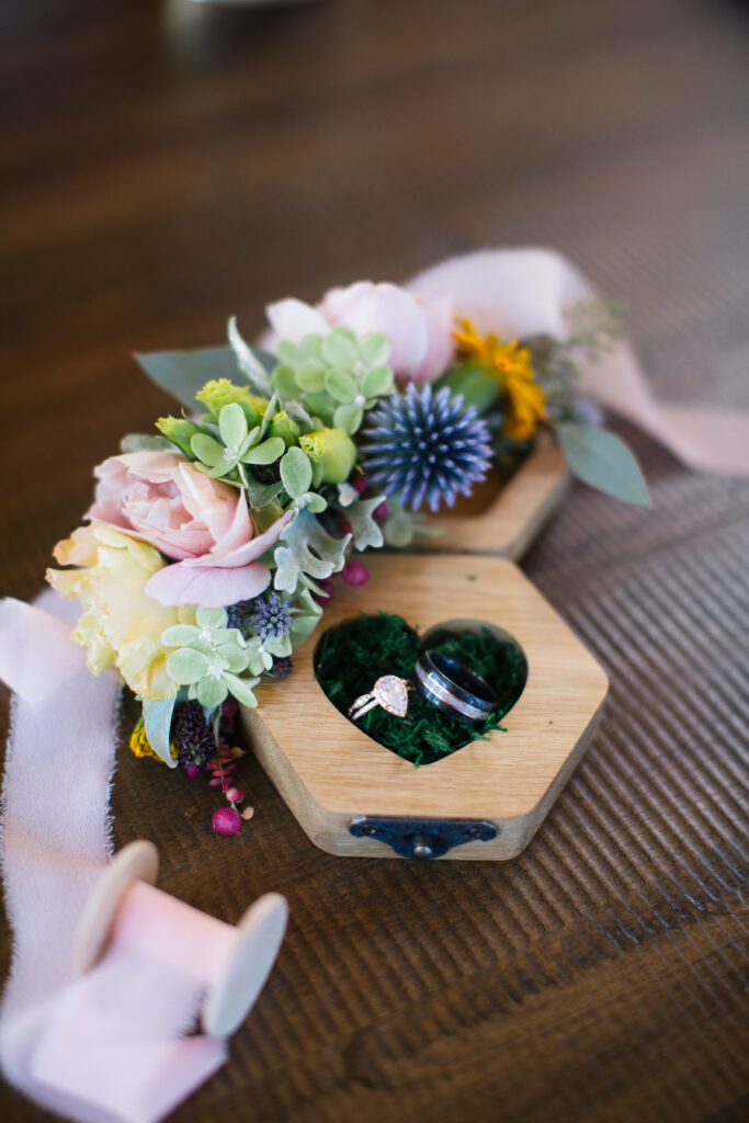 Wood ring box adorned with flowers on a wood table with a pink ribbon surrounding the box of rings.