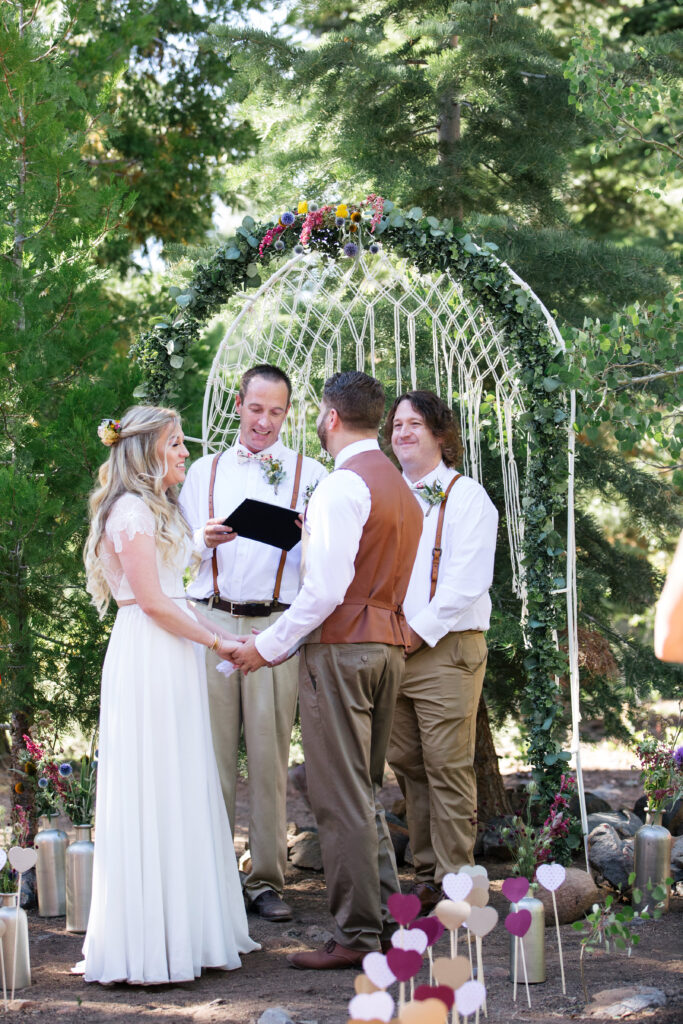 A wedding ceremony in a pine grove with two men in matching white shirts, suspenders and slacks as the officiants, bride in a two-piece white dress and groom in a tan vest, white shirt and tan slacks.