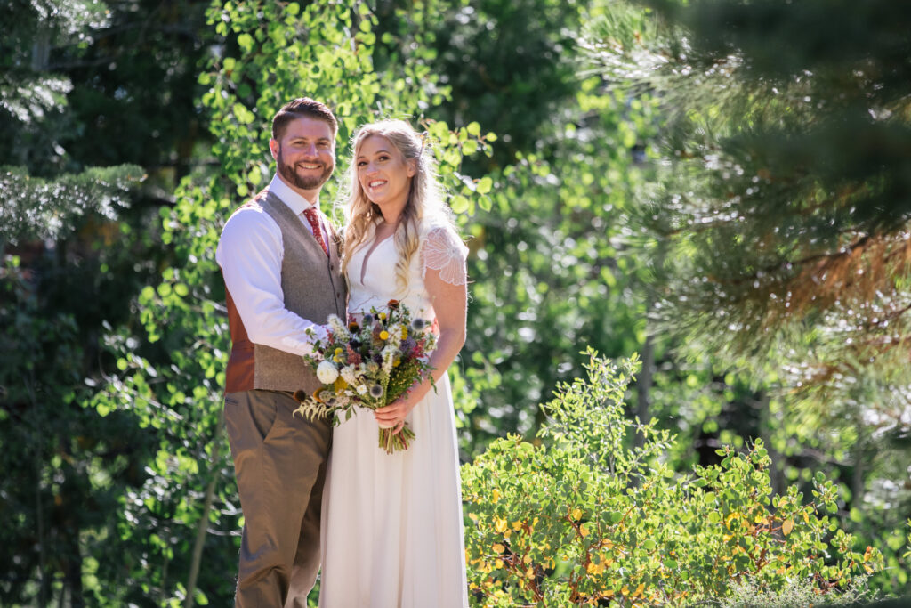 Woman in a white two-piece dress holding a bouquet standing with a man in a tan suit within a forest, both smiling.