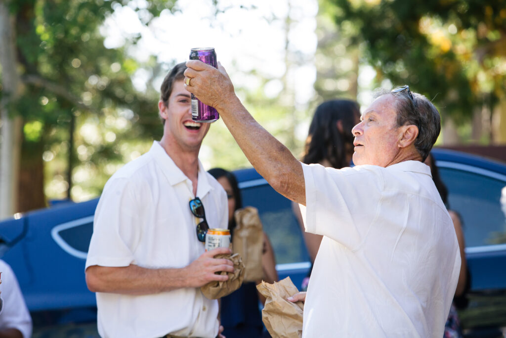 Man in a white shirt holding a beer can aloft while another man behind in laughs.