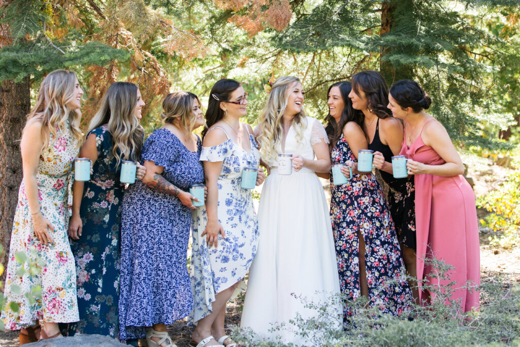 Group of women in floral dresses all smiling at the center woman in a white dress, all holding coffee mugs.
