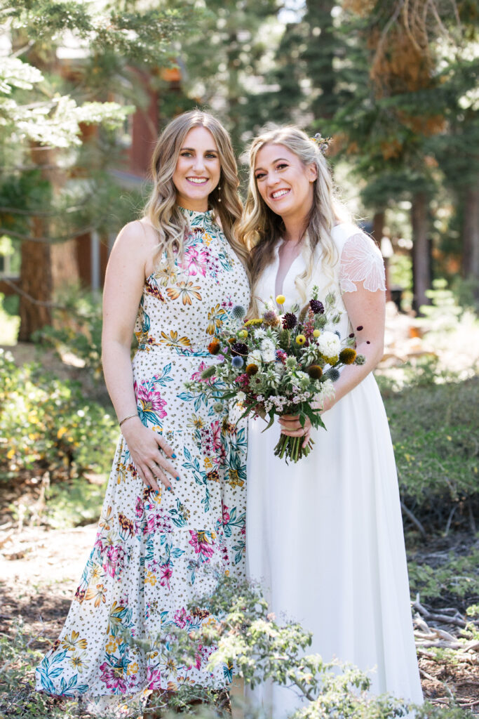 Two blonde women standing together, one in a floral dress and the other in a white gown holding a bouquet.
