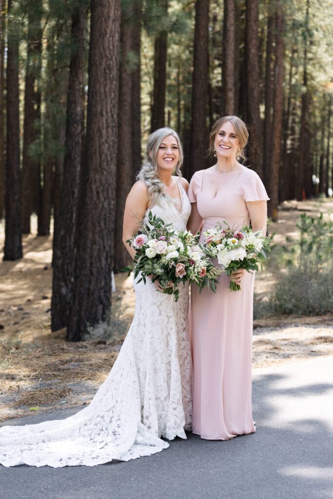 Woman in a white lace gown holding a bouquet of flowers standing next to a woman in a blush dress holding a bouquet of flowers with pine tress behind them.