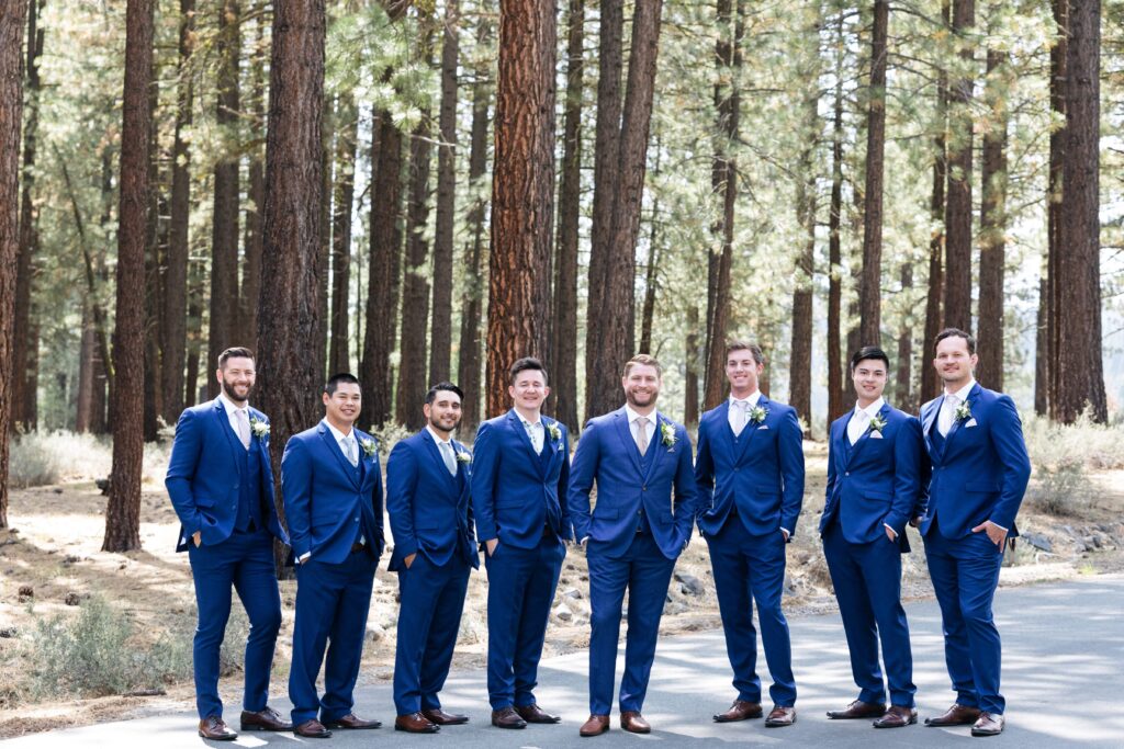 Group of men in dark blue suits and dark leather shoes standing with their hands in their pockets in front of pine trees.