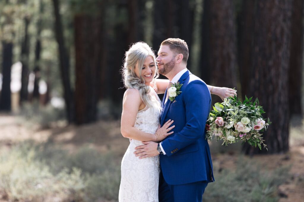 Woman in a white lace gown holding a bouquet of flowers over a man's shoulder while they embrace.