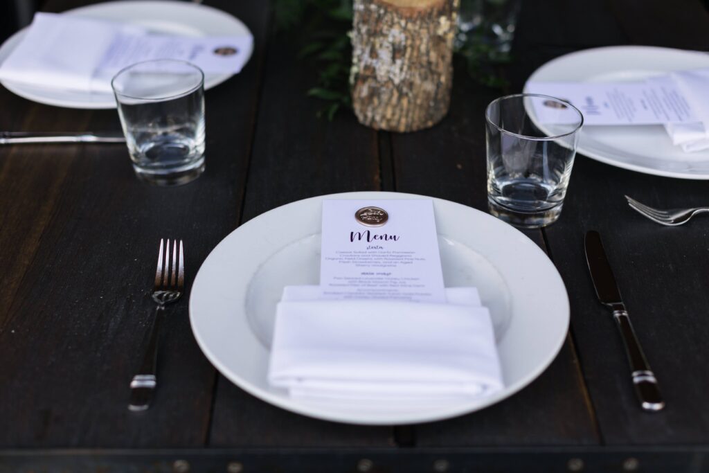 Table setting with a white pocket fold napkin holding a menu card atop a white plate, a low tumbler water glass, silver flatware, all on a wood table.