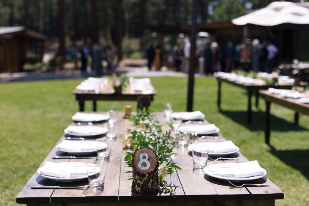 Wedding tables set with white plates and napkins, wood log table numbers, and greenery down the center of each table.