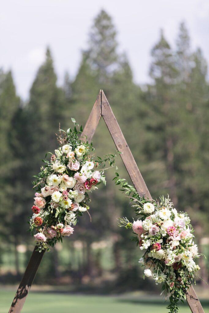 Wood triangle arch adorned with flowers in front of pine trees.