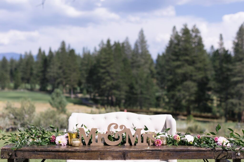 Wedding sweetheart table with a wood sign reading "Mr&Mrs" in front of a row of greenery and pink flowers atop a wood table in front of a white settee with a backdrop of pine trees.