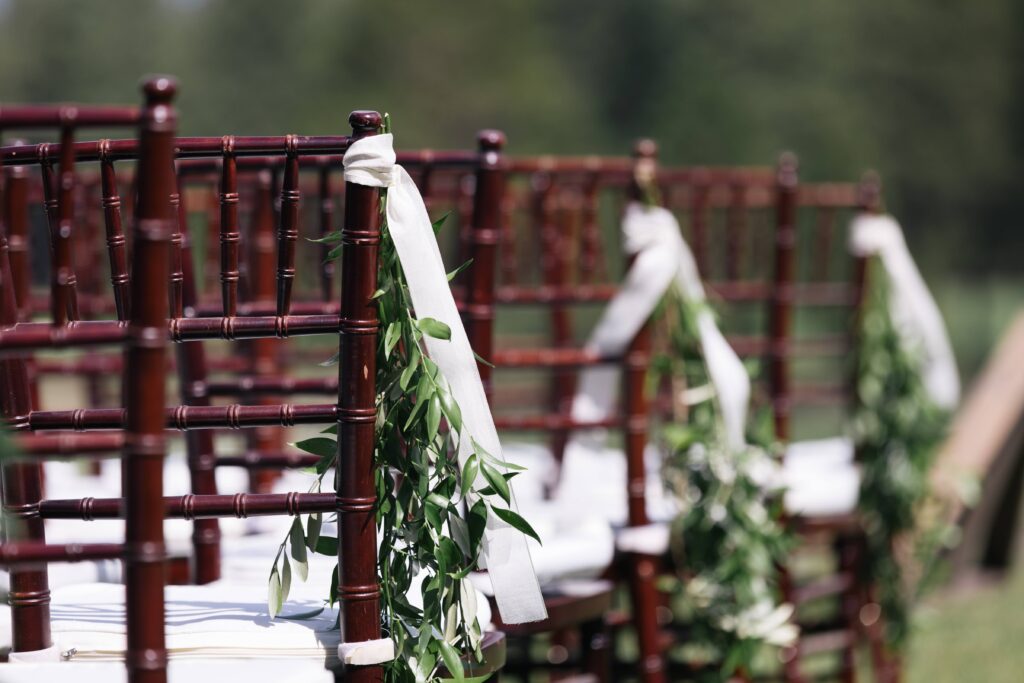 Dark brown chivari chairs with white pads aligned in neat rows, every other row starting chair adorned with a white ribbon and greenery.