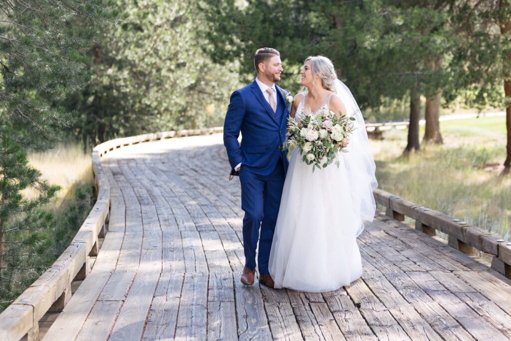 Wedding couple smile at each other while walking down a wooden walkway through pine trees.