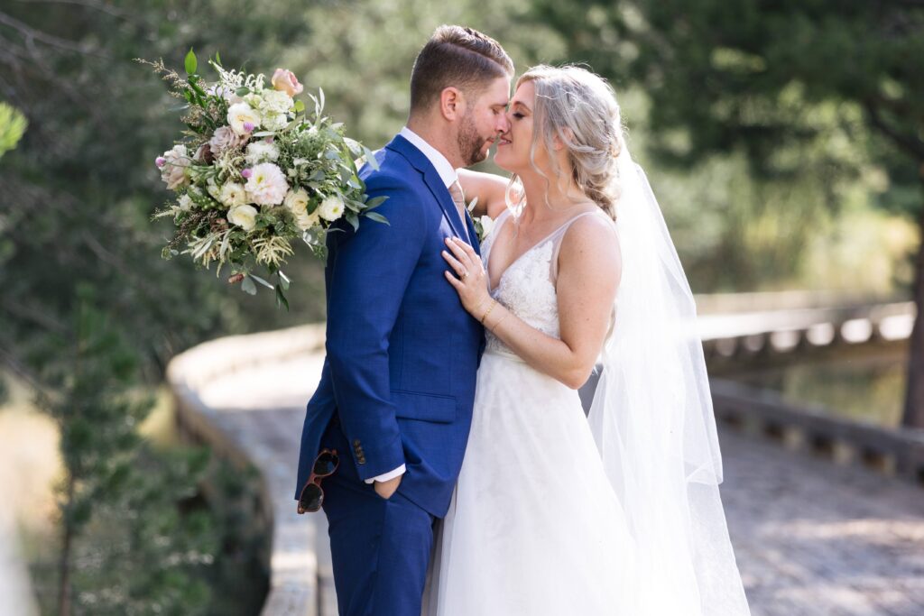 Woman in a white lace gown and veil holding a bouquet of flowers leans into a man in a dark blue suit for an embrace.