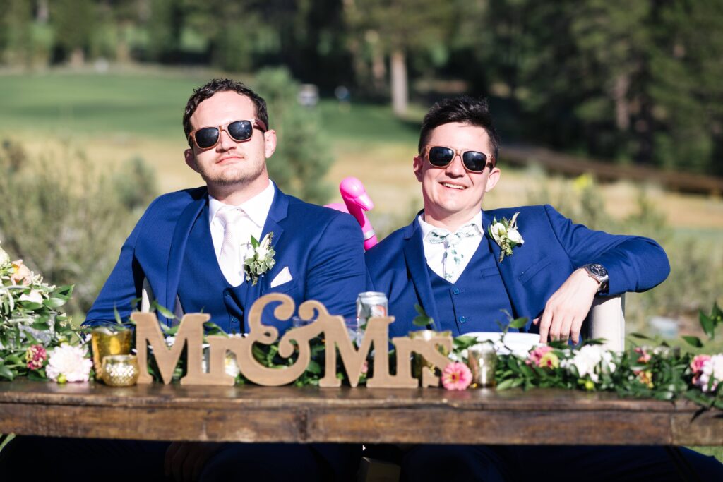 Two men in matching dark blue suits wearing sunglasses sit behind a table with a sign reading "Mr&Mrs".