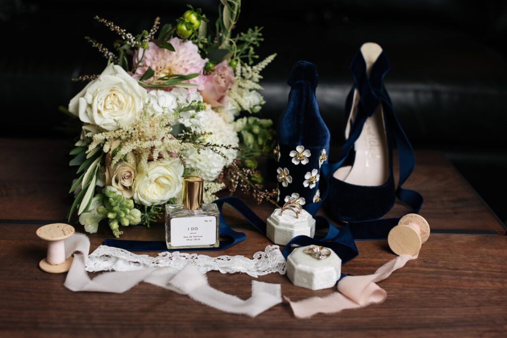 A bouquet of white and pink flowers, perfume bottle reading "I Do" on the label, dark blue velvet heels with white metal daisies, spools of pink ribbon, white velvet ring box, and white lace garter.