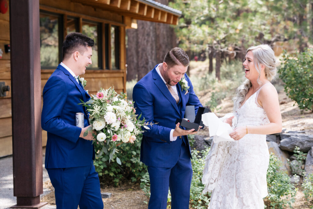 A man in a blue suit staring in awe at an opened black watch box, a woman in a white lace gown laughs to his side, and another man in a blue suit smiles on his other side holding a flower bouquet.