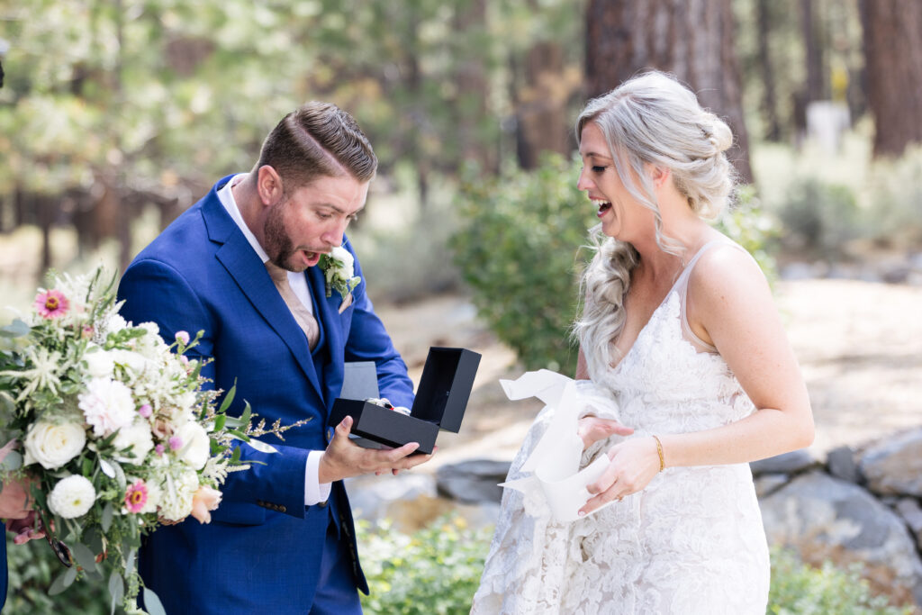 A man in a blue suit staring in awe at an opened black watch box and a woman in a white lace gown laughs to his side.