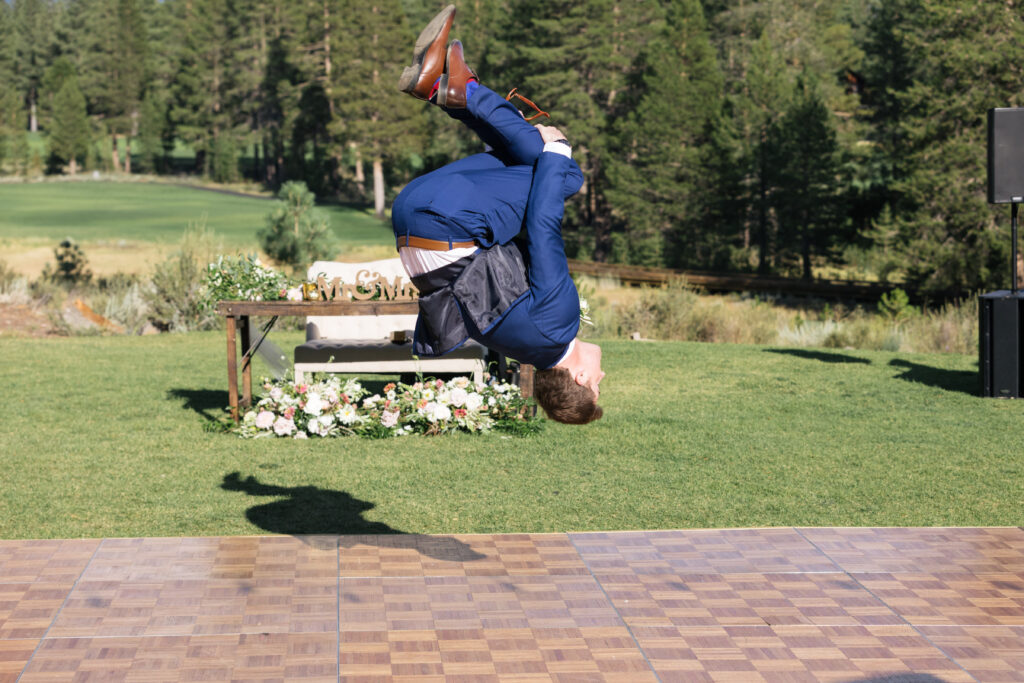 A man in a blue suit doing a backflip over a wood dance floor outside amongst pine trees.