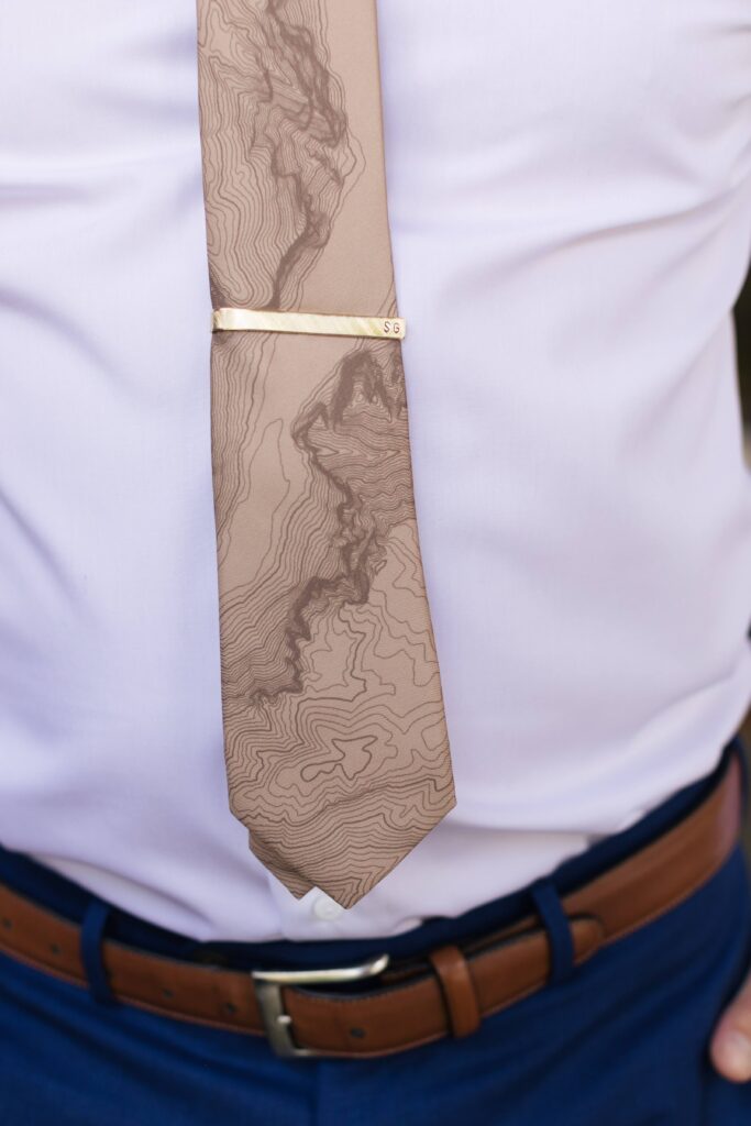 Close up of a topography printed tan necktie atop a white button-down shirt above a leather belt holding dark blue slacks.