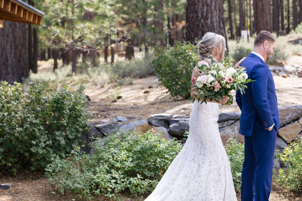 Woman in white lace gown holding a bouquet of flowers approaches a man in a dark blue suit with his back to her.