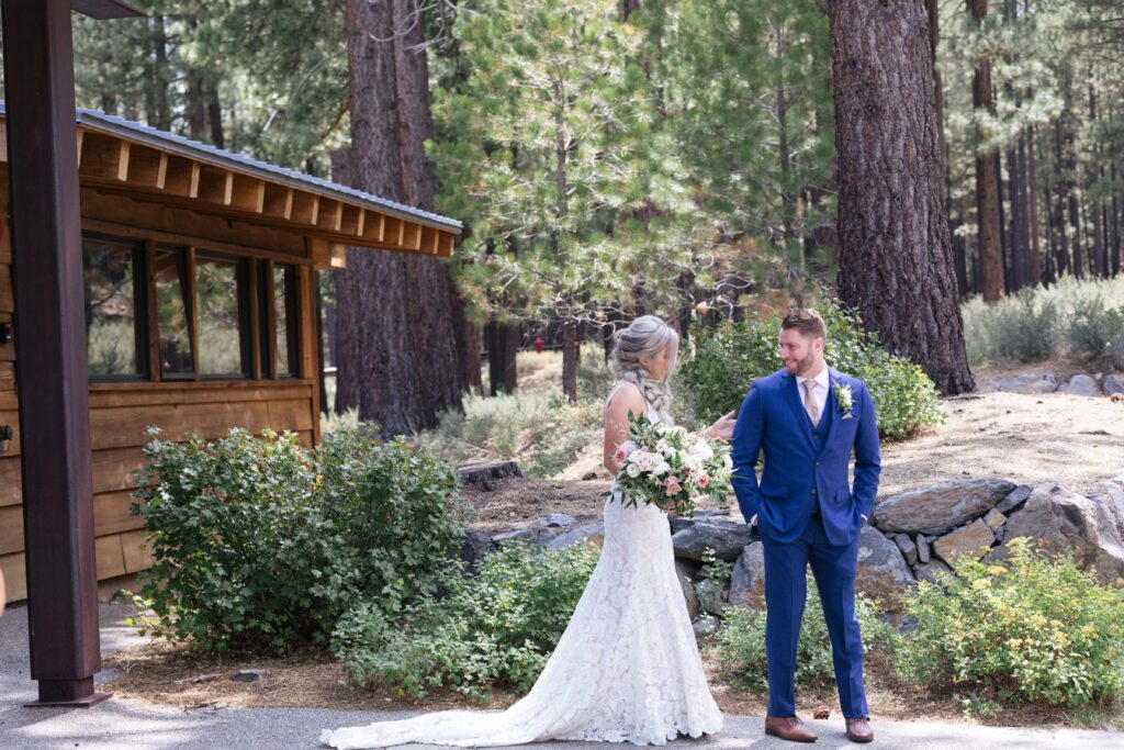 Woman in a white lace gown holding a bouquet of flowers stands before a man in a dark blue suit within a grove of evergreen trees.