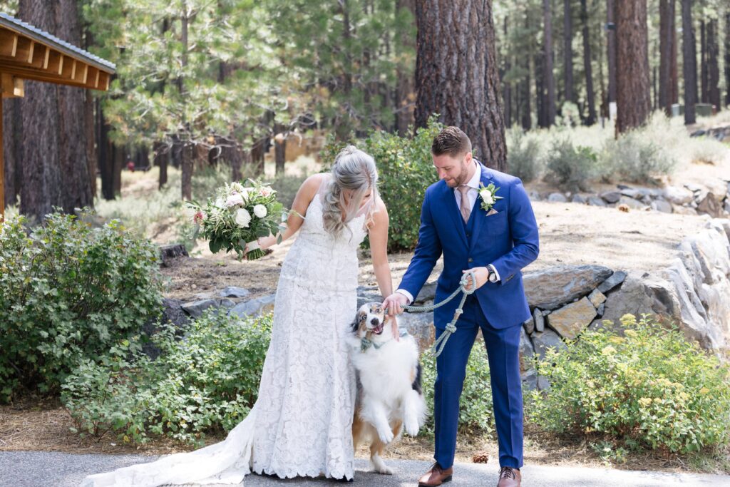 Woman in a white lace gown holding a bouquet of flowers pets an Australian shepherd being held by a man in a dark blue suit within a forest of pines.