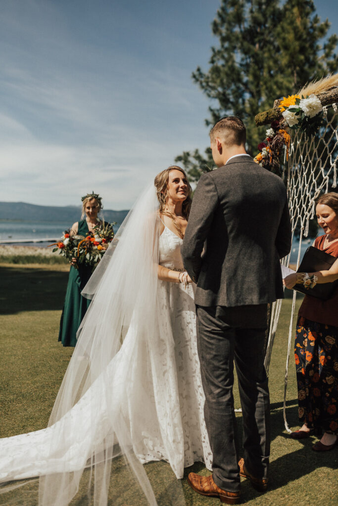 Wedding ceremony with the bride in a white gown and veil, groom in a grey suit with tan cowboy boots, maid of honor in a green dress, and officiant in a rust top and floral skirt under a oak limb arbor all in front of a lake.