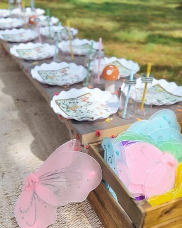 A low outdoor table covered with a transparent floral tablecloth, set atop a tan rug. The table is arranged with white plates, fairy-themed paper napkins, plastic milk bottles with colorful striped straws, toadstool decorations, and a wooden crate filled with glittering fairy wings in various colors at the head of the table.