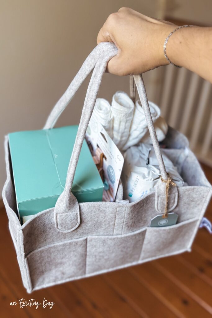 Hand holding a grey diaper caddy, with a little teddy bear keychain, filled with burp cloths, wipes, and other baby related items.