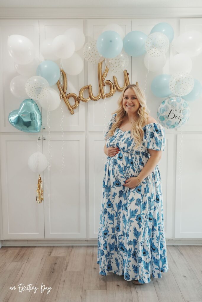 Blonde woman in a white and blue floral dress standing in front of white cabinets decorated with white and blue balloons with a sign reading "baby".