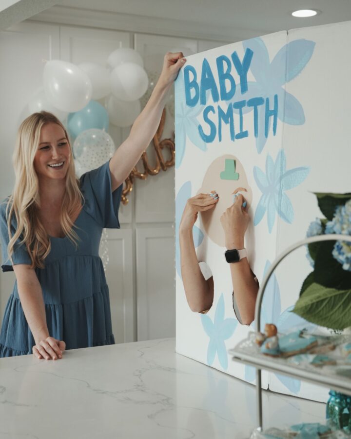 Blonde woman standing next to a marble countertop holding a poster board with blue flowers and a blank face painted on the front, and two holes cutout towards the base with two arms reaching through touching the poster face, with the phrase "baby smith" written across the top of the poster.