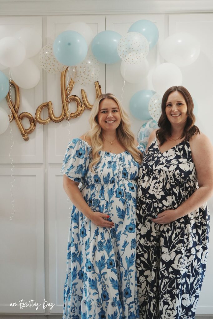 Two women, one blonde in a white and blue floral dress and the other brunette in a dark blue and white floral dress, standing in front of white cabinets decorated with white and blue balloons with a sign reading "baby".