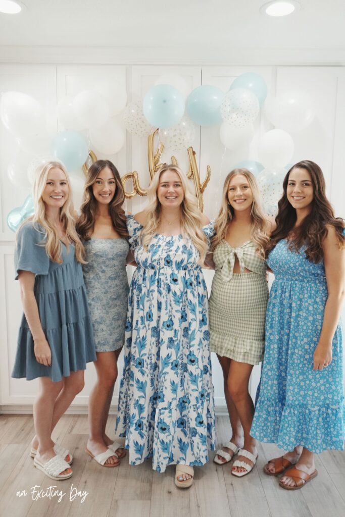Group of blonde and brunette women in spring blue, white and green dresses, standing in front of white cabinets decorated with white and blue balloons with a sign reading "baby".