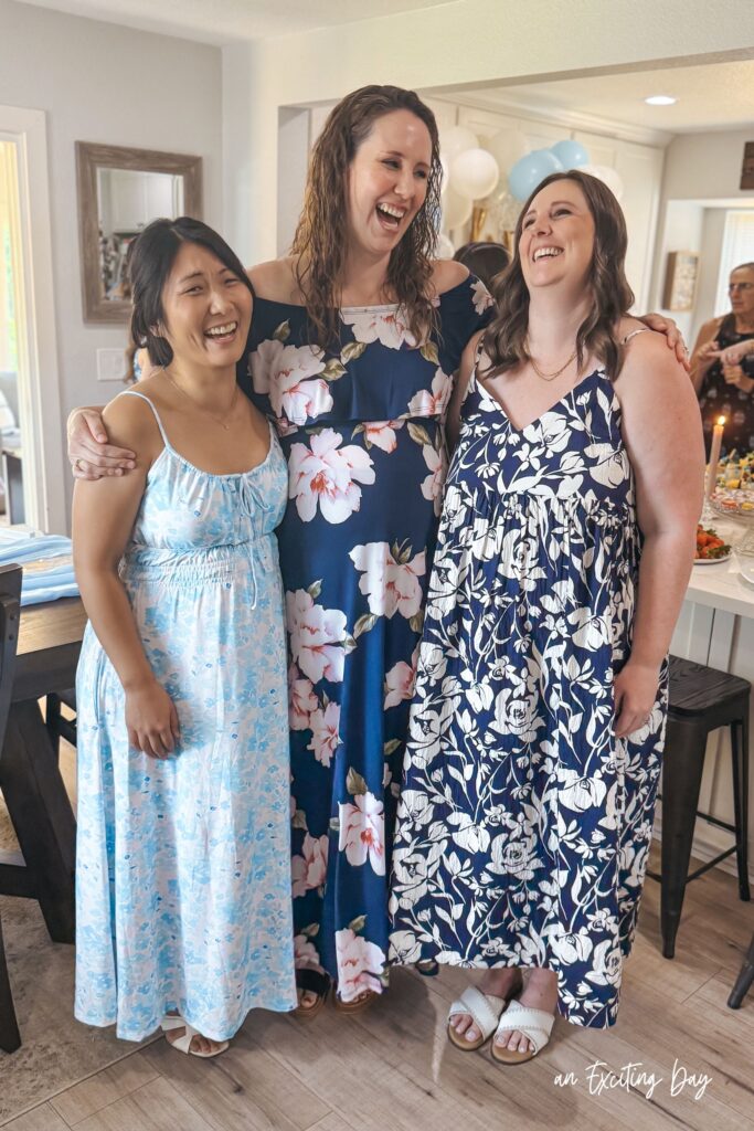 Group of brunette women in blue floral dresses smiling and laughing together inside a home with white walls and light wood floors.