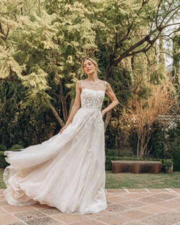 Woman standing in a courtyard wearing a wedding dress with thin straps and 3D flower petals cascading down the bodice.