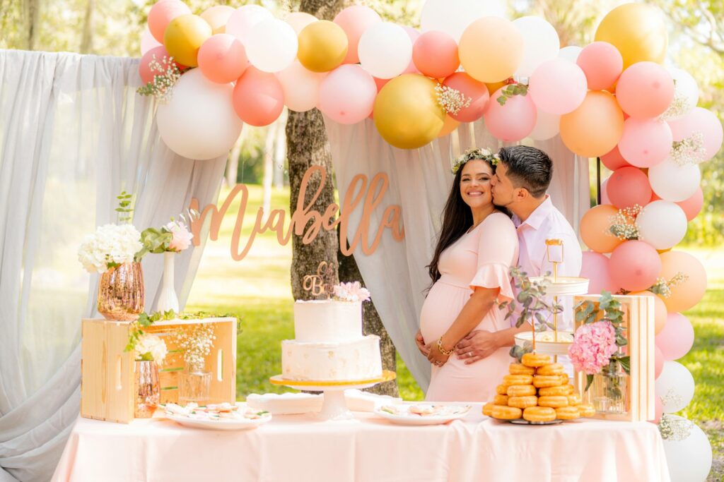 Man kissing a woman on the cheek as she embraces her pregnant belly under pink, white and hold balloons in front of white draped linens and behind a table with treats like cake, donuts and cookies.