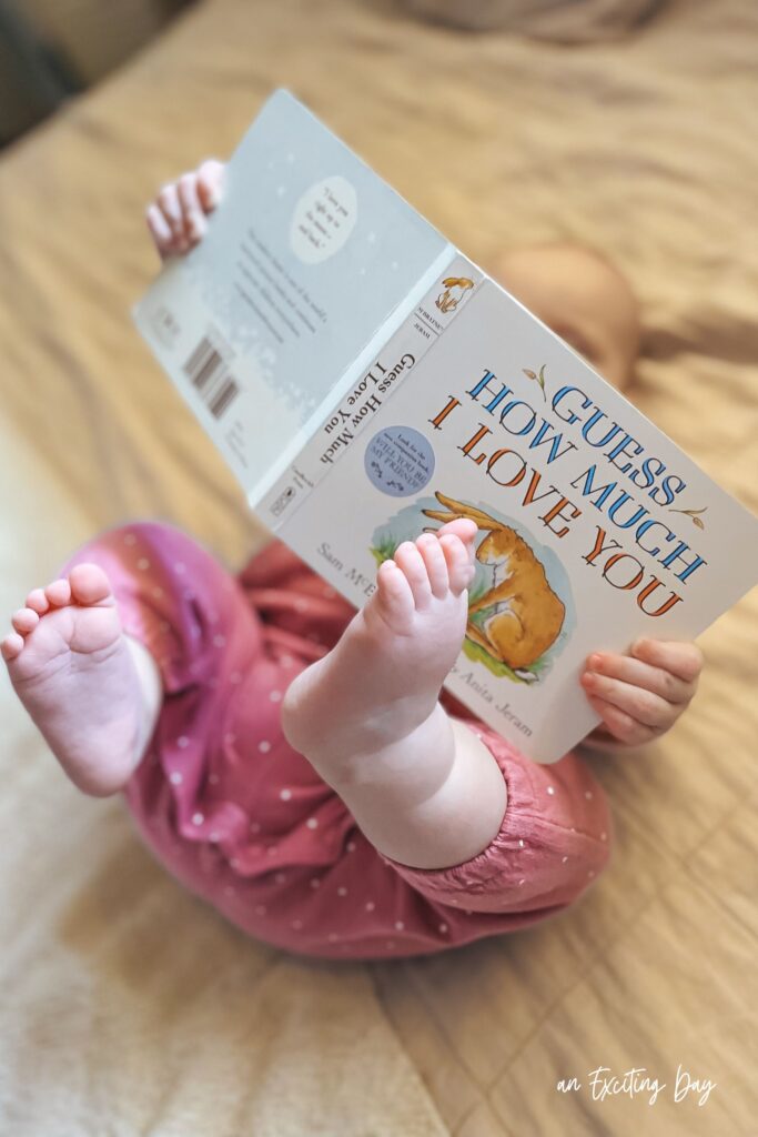 Baby laying on their back with their feet in the air holding a book above them that says "guess how much I love you."