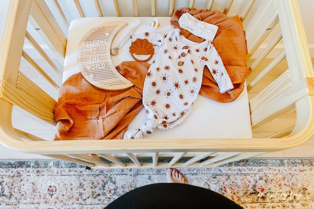 Birdseye view of a mini wooden crib with a wooden moon sign laying in the crib that reads "welcome to the world" along with a rust colored blanket and a baby sleeper that's white with rust colored suns.
