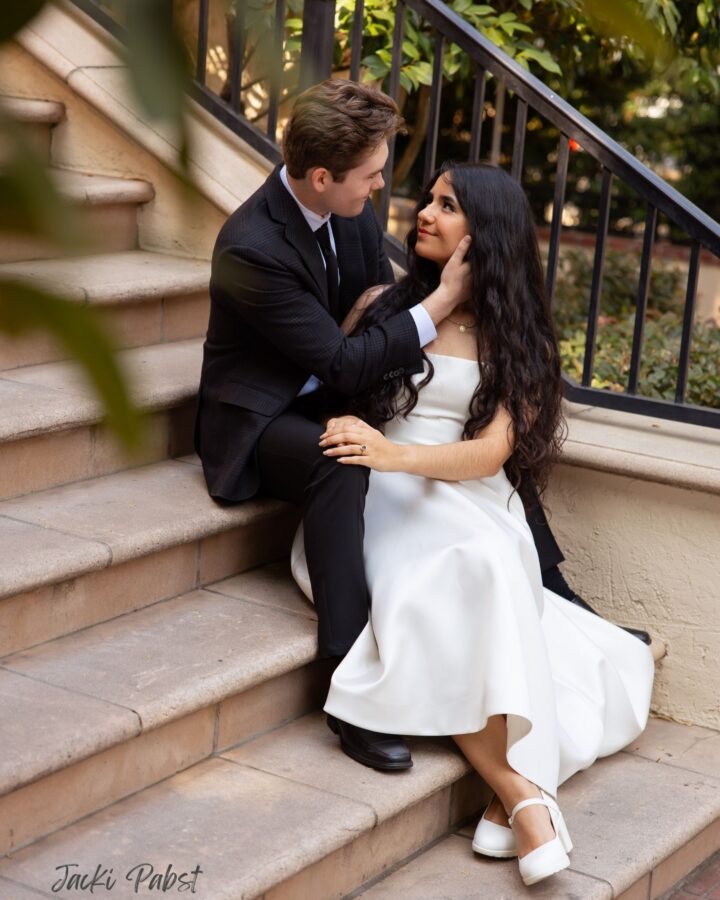 Couple seated on stone stairs of a building looking at each other in an embrace with the man seated one stair higher than the woman and wearing a black suit, while the woman with black hair wears a white dress and white heels.