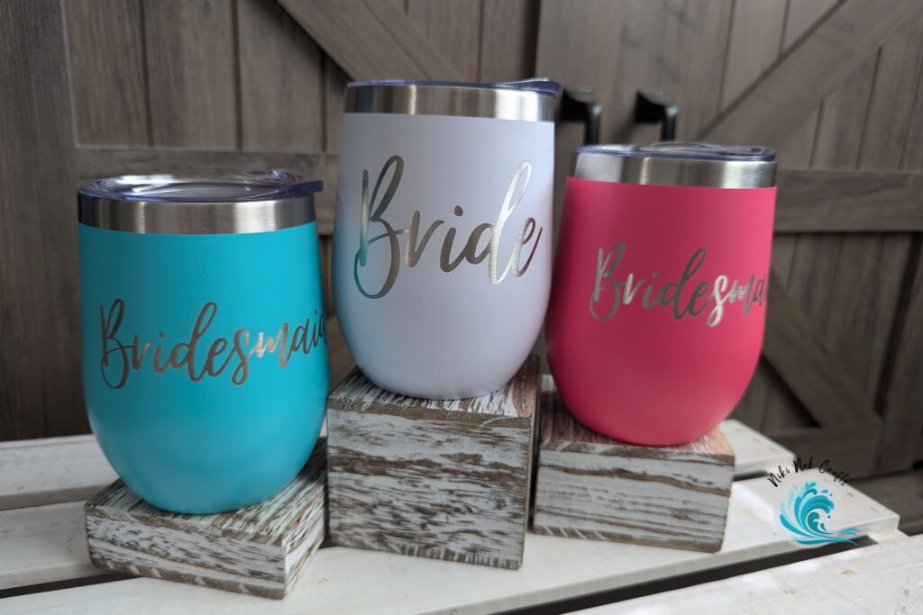 Metal wine tumblers engraved with "Bridesmaid" on the teal metal cup, "Bride" on the white metal cup, and "Bridesmaid" on the pink metal cup.