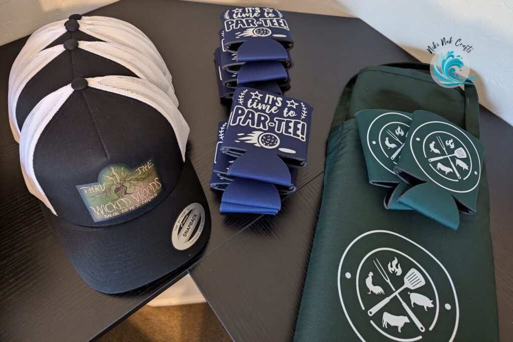 A stack of trucker ball caps, blue koozies and green apron with matching green koozies.