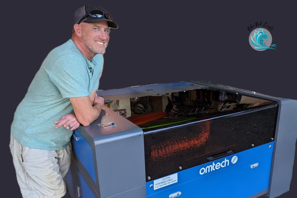A smiling man wearing a black ball cap with sunglasses perched on top, light teal shirt, and tan cargo shorts leaning on an "omtech" laser machine.