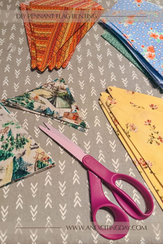 Sewing project scene with stacks of different printed fabrics spread across a table and fabric shears angled between two pieces of fabric as if it had just cut a triangle off the other piece of fabric. Overlay text reads "diy pennant flag bunting: and "www.anexcitingday.com".