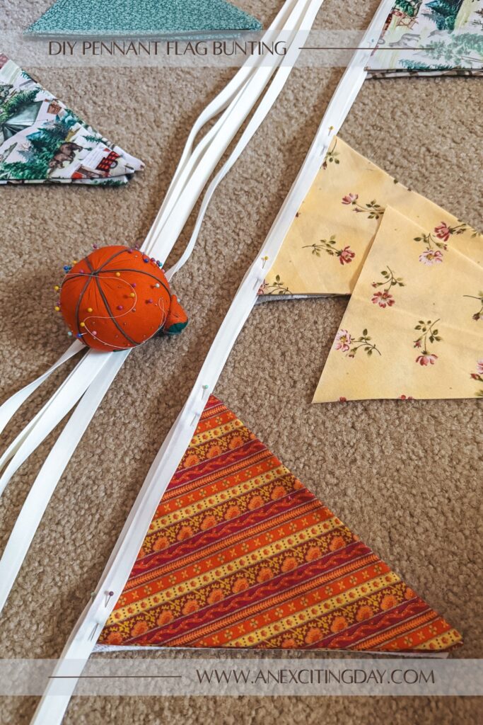 Sewing project scene with stacks of fabric triangles in orange stripe, yellow floral and camping prints, with white bias tape near the triangle shaped fabric pieces and a pin cushion shaped like a tomato. Overlay text reads "diy pennant flag bunting: and "www.anexcitingday.com".