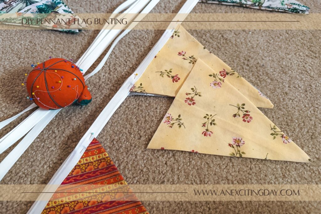 Sewing project scene with stacks of fabric triangles in orange stripe, yellow floral and camping prints, with white bias tape near the triangle shaped fabric pieces and a pin cushion shaped like a tomato. Overlay text reads "diy pennant flag bunting: and "www.anexcitingday.com".