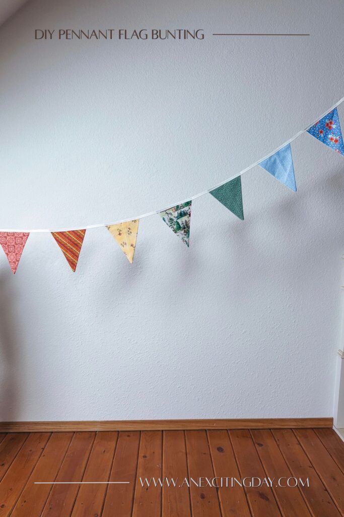 Colorful pennant flag bunting with red, orange stripe, yellow floral, green print, green floral, and blue floral fabrics strung above a pine wood floor. Overlay text reads "diy pennant flag bunting: and "www.anexcitingday.com".