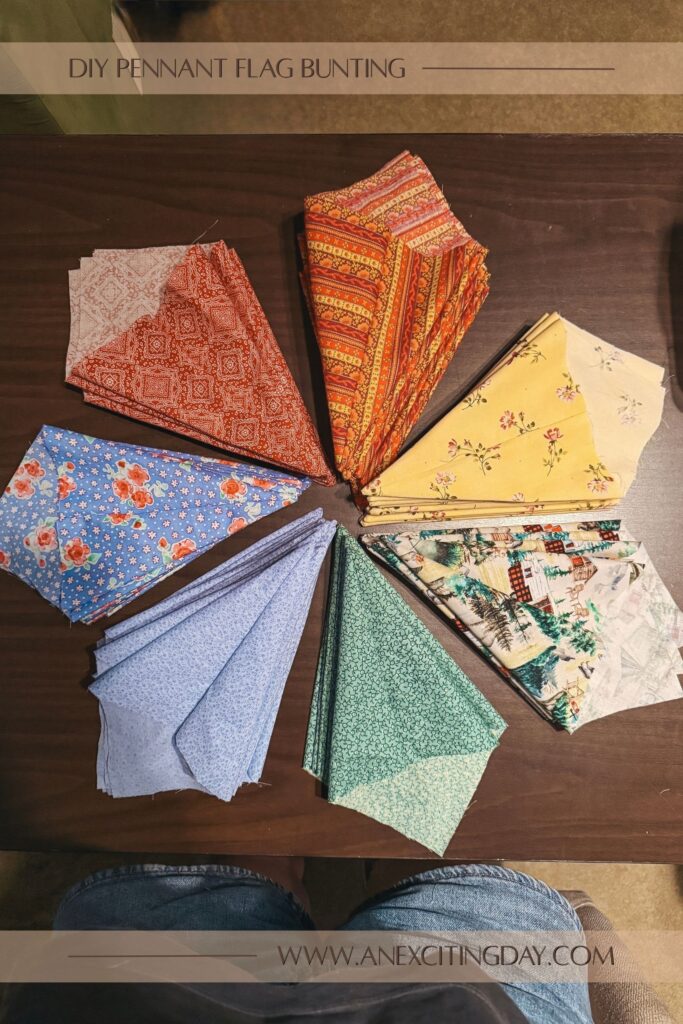 Seven stacks of sewn fabric, each a different color of the rainbow, in the shape of kites all arranged with their points angled at a center point of a circle atop a desk. Overlay text reads "diy pennant flag bunting: and "www.anexcitingday.com".